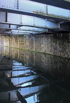 old steel girders reflected reflected in the water of a dark canal under a bridge