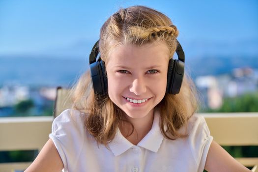 Head shot, portrait of child girl looking outdoors at camera. Preteen in headphones, smiling blonde, face close up, young female student, blue sunny sky background