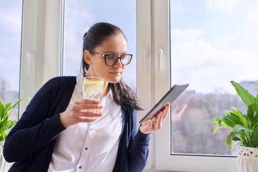 Adult woman drinks water with lemon, female in glasses with digital tablet, at home near window