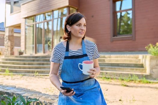 Portrait of middle aged woman relaxing outdoors in backyard. Female in garden apron with mug of tea and smartphone in her hands, looking away, spring garden background. 40s people, lifestyle