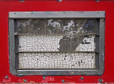 an old scruffy caravan window with steel frame and slatted windows with cracked old white paint with a red surround with rivets