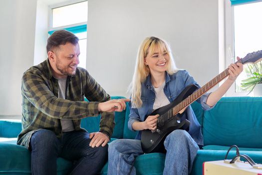 Father teaching teen daughter to play electric guitar. Family seated on sofa in living room. Creativity, adolescence, family, relationship, father and teenager, dad and teenage daughter, father's day