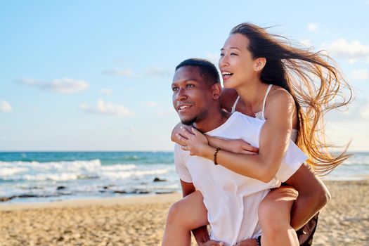 Young beautiful having fun couple on sea background. Multiracial couple, asian woman on back of african american man, vacation together, relationship dating happiness, happy lovers moments, copy space
