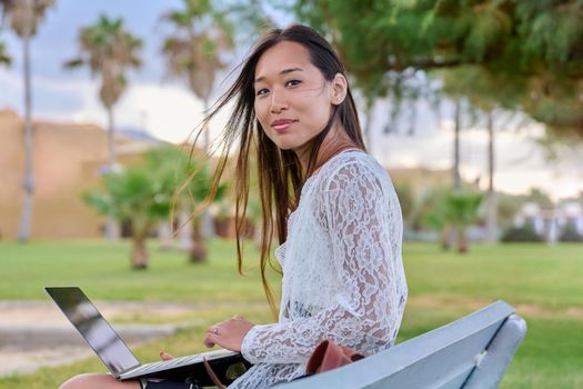 Young beautiful woman outdoors with laptop, tropical park background. Asian female sitting on bench, working, studying remotely. Freelance e-learning, online technologies, education, business concept