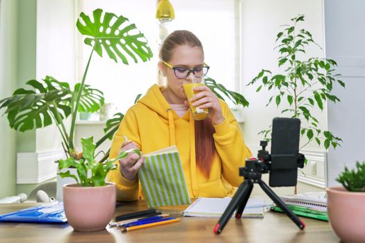 Teenage girl looking at webcam using smartphone for an online lesson, reading textbook book. Student 12, 13 years old, studying remotely at home, e-learning, virtual lessons, distance learning