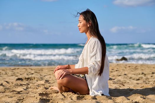 Young beautiful woman in lotus position meditating on the beach. Relaxing resting Asian female with closed eyes sitting on sand, scenic seascape background. Lifestyle, health, beauty, nature, people