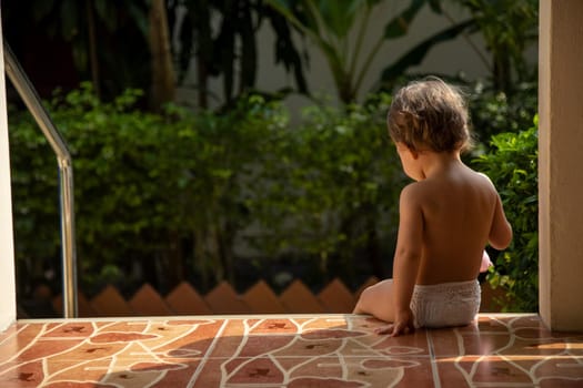 a charming toddler sits on the steps of a house on green garden background in the sunlight . back view