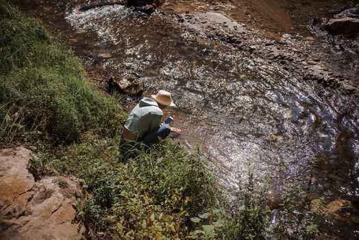 Woman with hat, back profile sitting and taking pictures with the phone of shining stones in the running water of a mountain river, in summer with green foliage, stones and flowing stream.