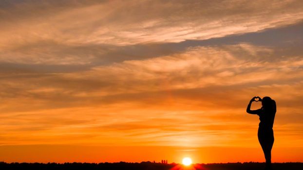 Woman in love making a heart symbol from fingers in a fire sky background. Symbol of love and beautiful things. Silhouette of a happy woman.