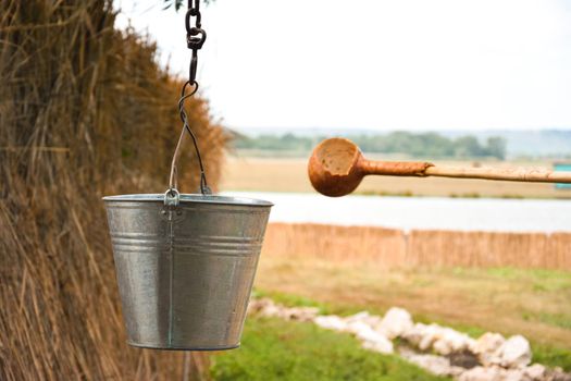 A water fountain with an old bucket and a makeshift mug made of pumpkin. Reed fence in background. Traditions and lifestyle.