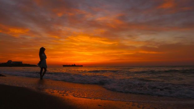 Woman with long hair and summer dress looking at the sea at sunrise. Enjoy the sea breeze in the morning. Colorful summer landscape. A big ship on bacround