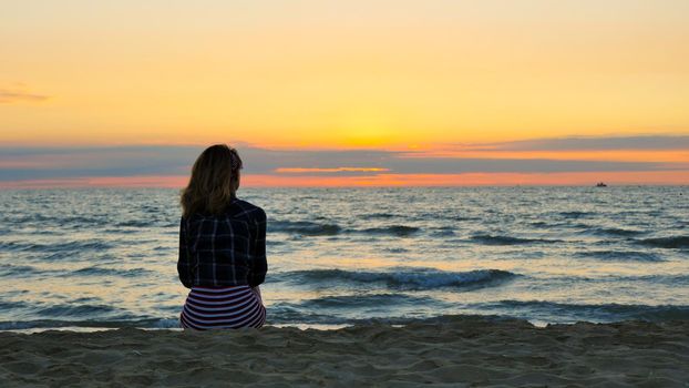 Blonde girl relaxing on the beach and watching the sunset. Moment of meditation. Sea, waves and orange sky in the background