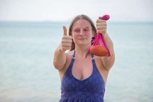 woman holding mesh bag with fruit and showing thumbs up. on the background is sea or ocean.