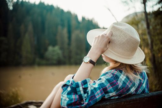 Rear view of a young woman in a denim shirt and hat, sitting on a bench, admires the lake landscape, the pine forest and the relief of the mountains in the background