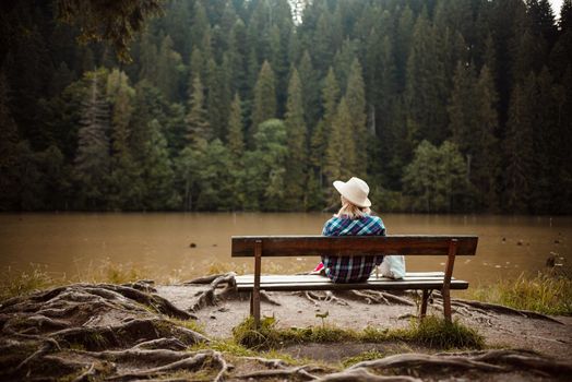 Rear view of a young woman in a denim shirt and hat, sitting on a bench next to a tree with many roots, admires the lake landscape, the pine forest and the relief of the mountains in the background, in Red Lake, Eastern Carpathians, Romania