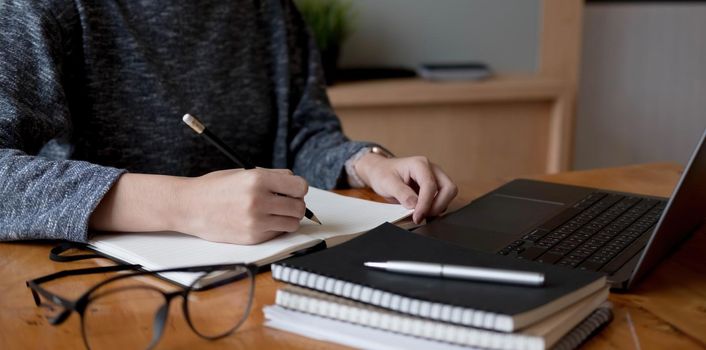 Cropped photo hand of woman writing making list taking notes in notepad working or learning online with laptop at home.