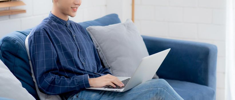 Young asian businessman smile and work from home with laptop computer online to internet on sofa in living room, freelance man using notebook on couch with comfort, new normal, lifestyle concept.