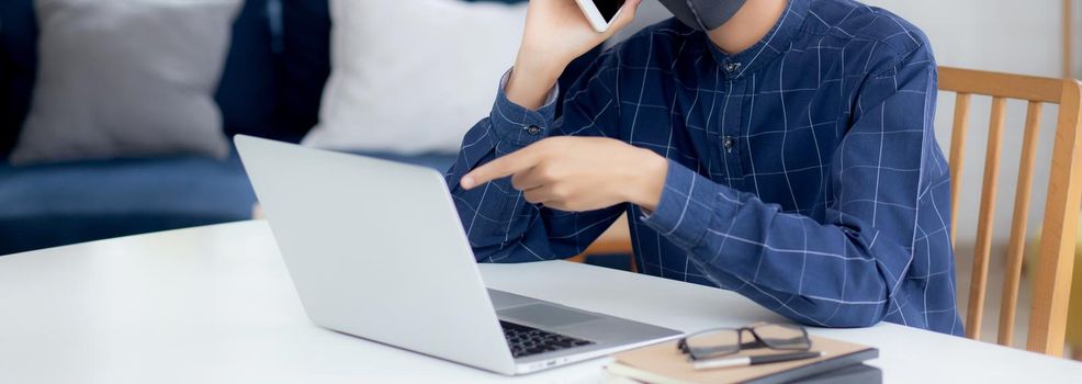 Young asian business man in face mask talking smartphone and work on laptop computer for protect covid-19, businessman quarantine work from home with notebook, social distancing and stay home.