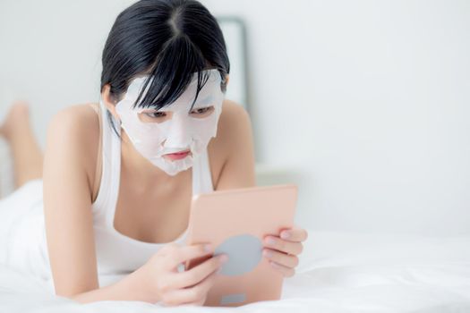 Beautiful young asian woman with sheet facial mask lying watching tablet computer on bed at bedroom, beauty girl applying face mask for skincare of wrinkle at home, skin care, health and wellness.