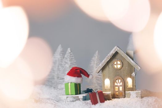 Magic Christmas card with toy wooden house in winter forest and gifts, bokeh lights around