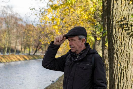 Elderly pensioner stands on beautiful autumn day in city park and rests while walking. The man holds his cap by the visor. Selective focus.