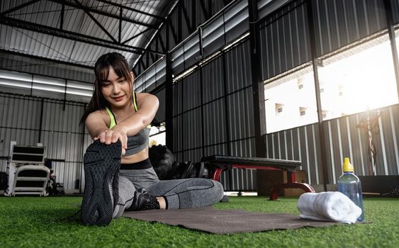 Young asian female in stylish and comfy sportswear stretching her legs on the fitness floor. exercise and sport concept. Fit woman doing stretching workout at gym.