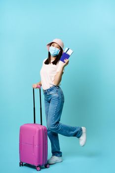 Full length of happy asian girl tourist, woman on vacation standing with suitcase, showing tickets and passport, travelling abroad, blue background.