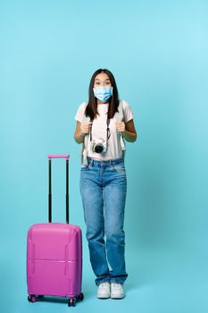 Enthusiastic girl traveller, asian tourist with backpack and suitcase, wearing medical face mask, excited about journey, vacation trip, standing over blue background.