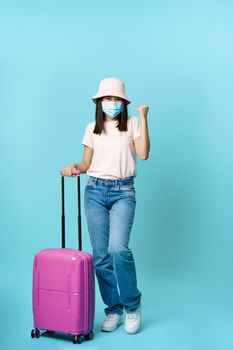 Enthusiastic girl, asian tourist in medical mask, holding suitcase, full length shot, travelling abroad on vacation during covid-19, blue background.