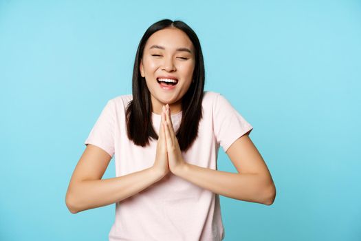 Smiling happy asian woman shows namaste, beg gesture and dreaming of something, making wish, high hopes, standing over blue background.