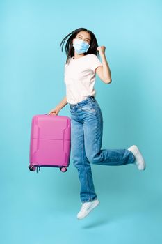 Full length of happy asia female tourist, dancing and jumping with suitcase in medical face mask, excited about vacation, standing over blue background.