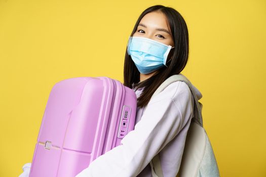 Close up of happy asian girl tourist in medical face mask, holding pink cute suitcase, going on vacation, travelling during pandemic, yellow background.