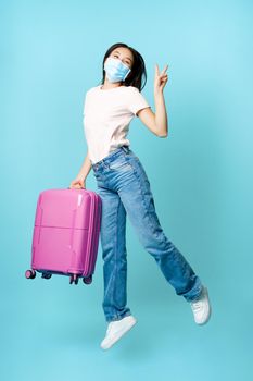Full length of happy asian woman jumping from joy with suitcase, wearing medical face mask from covid, excited about vacation abroad, tourism and healthcare concept.