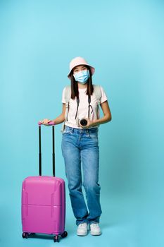 Smiling korean girl on vacation during pandemic. Standing with suitcase and photo camera, wearing medical face mask, blue background.