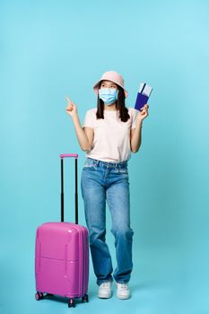 Smiling asian traveller, tourist girl in face mask, standing near suitcase with two tickets and passport, pointing aside thoughtful, choosing vacation destination, blue background.