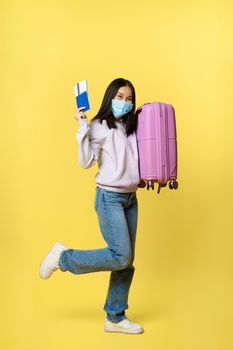 Full length shot cheerful asian female tourist, girl in medical face mask dancing with suitcase and passport with tickets, going abroad on vacation during pandemic.