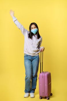 Full length shot happy asian woman going on vacation, tourist with suitcase posing cheerful, wearing medical face mask, yellow background.