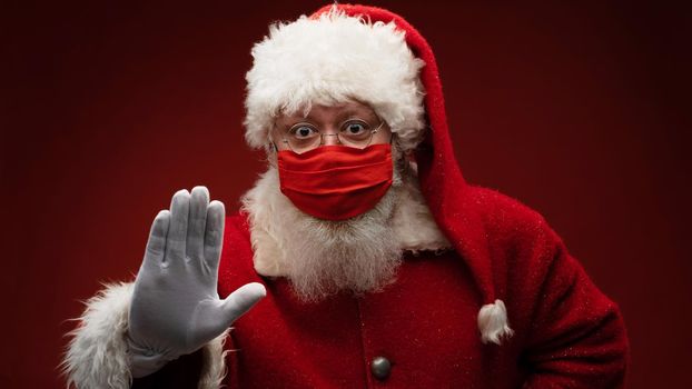 Real Santa Claus on red background, wearing protective mask and with an open hand in front making a pandemic stop sign. Christmas with social distance. Covid-19