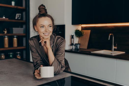 Relaxed positive young woman in silk cozy pajama having cup of coffee in stylish kitchen interior enjoying leisure morning time at home, looking aside with smile while starting new beautiful day