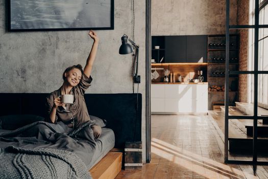Energy drink. Young beautiful positive european woman sitting on bed stretching with her hand up while holding cup of coffee with another hand, trying to wake up before going to shower in morning