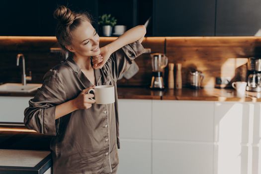 Young pretty relaxed woman in comfy pajama stretching from sleep early in morning while holding cup of coffee in her hand, standing in stylish kitchen interior. Leisure time on average day at home