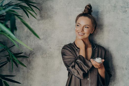 Young beautiful woman with glowing healthy skin broadly smiling while holding moisturizing face cream in her hand during morning cosmetic routine procedure at home, isolated over concrete wall