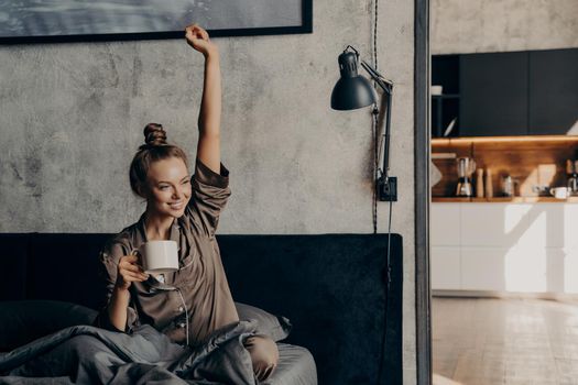 Happy life. Attractive young happy female having morning coffee in bed after waking up, dressed in satin pajama, stretching her hand up and enjoying new day while relaxing on weekend at home