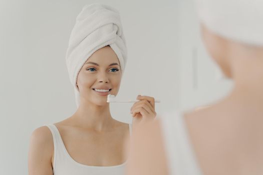 Portrait of young healthy beautiful woman with towel on head after shower having toothy smile while gently brushing her teeth, looking in mirror with pleasant facial expression. Dental health concept
