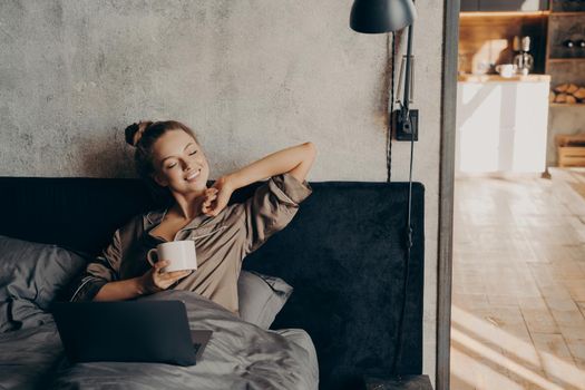 Happy beautiful female keeping eyes closed in satin pajama holding cup of coffee and enjoying morning sun, lying in bed with laptop after waking up in morning before starting working remotely at home