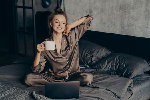 Relaxed female freelancer having cup of coffee stright after waking up in bed while checking new messages and emails on laptop, dressed in pajama. Woman with closed eyes ready to start remote work