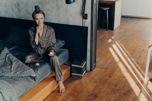 Skincare concept. Beautiful smiling young female applying moisturizing face cream during daily cosmetic routine procedures while sitting on bed after waking up in early morning in satin pajama