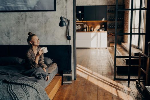 Young happy attractive girl sitting on bed in satin pajama in lotus position with cup of hot coffee in her hand while looking toward window smiling at morning sunlight, enjoying weekend at home