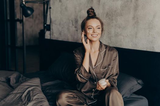 Young pretty european woman in silk pajama takes care of skin and body after waking up in morning at home while applying moisturizing face cream, getting ready for work. Beauty and skincare concept