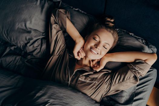 Good morning. View from above of young beautiful relaxed brunette female with closed eyes in satin pajamas waking up in her bed fully rested, happy woman stretching after night sleep